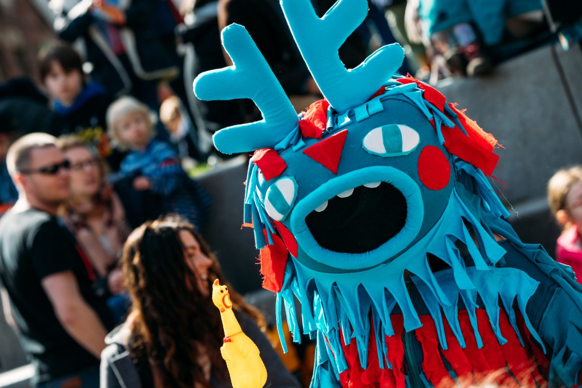A bright blue monster-style puppet costume with antler-like shapes on its head, multiple cartoon eyes, red fabric details and a large open mouth performs outdoors in front of a crowd. The puppet has long fringed fabric hanging from its face and body. In the foreground, a small yellow rubber chicken toy is held up toward the puppet, while adults and children watch in the blurred background at a daytime outdoor event.’