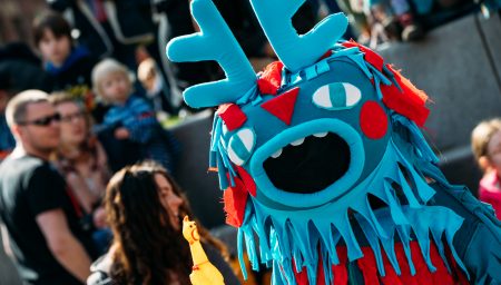 A bright blue monster-style puppet costume with antler-like shapes on its head, multiple cartoon eyes, red fabric details and a large open mouth performs outdoors in front of a crowd. The puppet has long fringed fabric hanging from its face and body. In the foreground, a small yellow rubber chicken toy is held up toward the puppet, while adults and children watch in the blurred background at a daytime outdoor event.