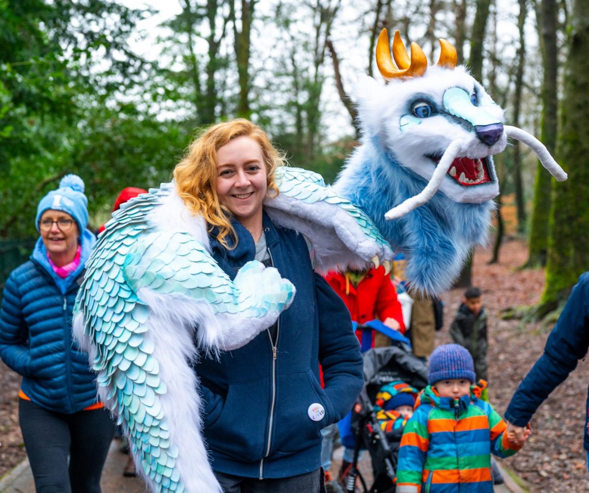 Person wearing a large dragon-like creature puppet around their shoulders during a woodland family event. The puppet has pale blue scales, white fur, golden horns, long whiskers and large expressive eyes.