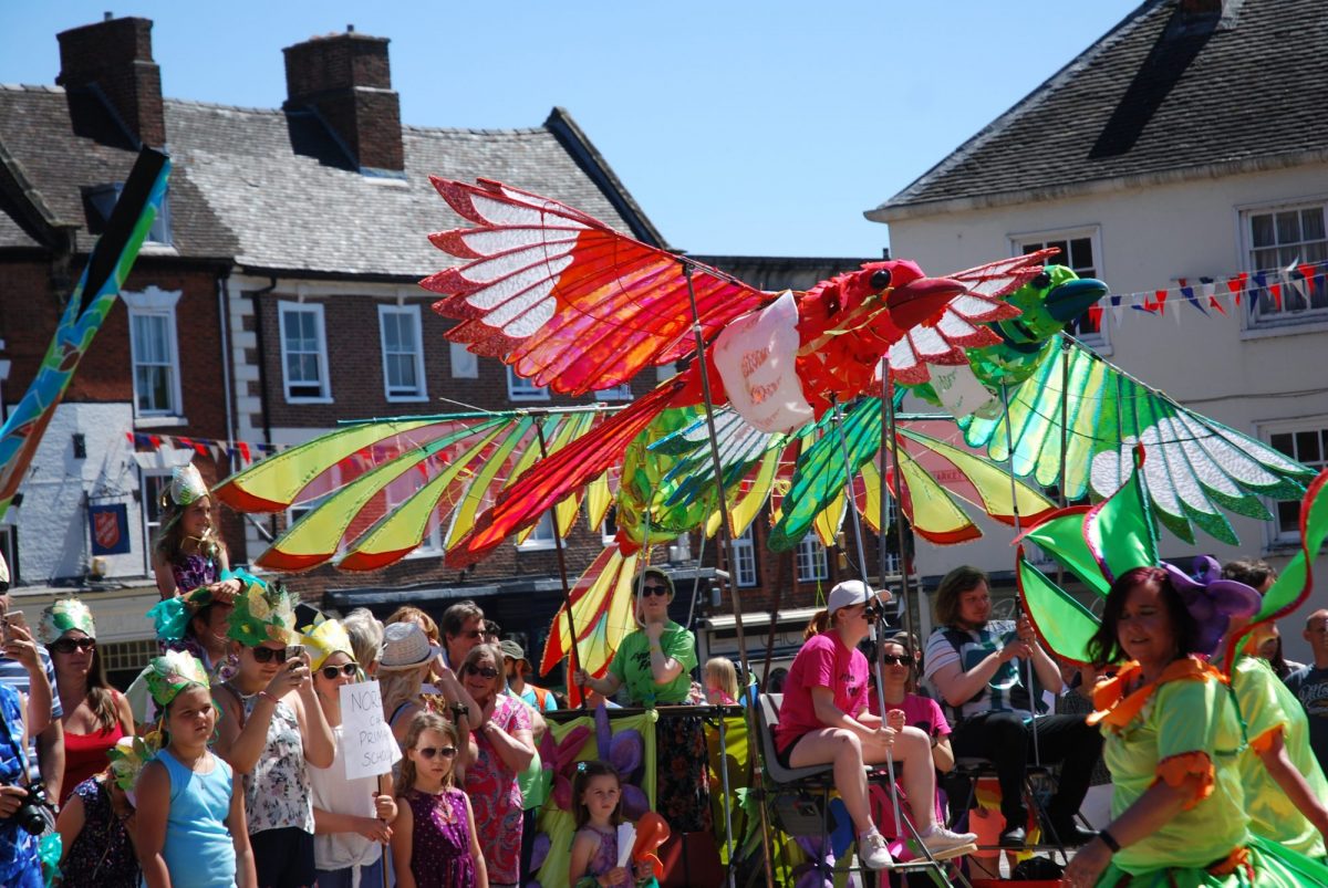 Two giant bird puppets with translucent red, green and yellow wings are carried above a crowd during a sunny outdoor parade.