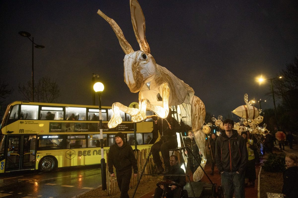 Night-time street parade featuring a giant illuminated hare puppet towering above a crowd of people. The glowing white hare has long ears and detailed woodgrain-style patterns across its body. Several puppeteers guide the structure from underneath and alongside it.
