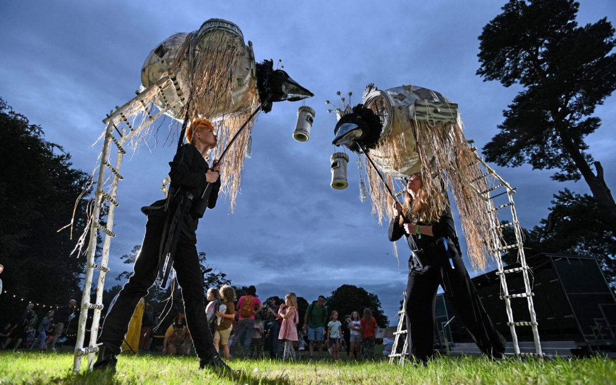 Two performers dressed in black operate giant bird-like puppets on long stilts during an outdoor evening event. The towering creatures have black beaks, pale woven bodies and long hanging strands resembling nests or feathers. Suspended lantern-like objects hang between them.