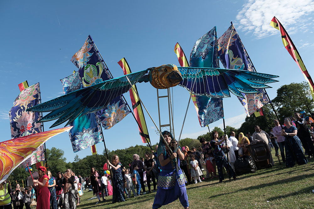 Large blue bird puppet with outstretched wings being carried through a festival crowd in a grassy park. The bird is decorated with turquoise, blue and gold patterns and is supported on poles by a performer in blue costume.