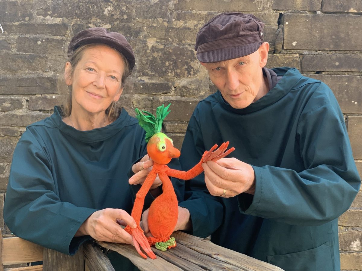Two performers wearing matching dark costumes and caps present a bright orange vegetable puppet with green leaves for hair.