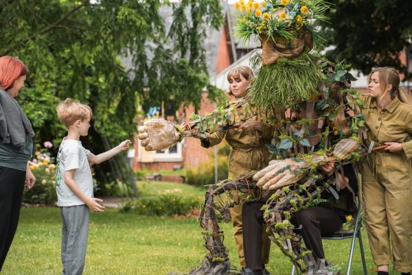 Large plant-like puppet made from branches, leaves, flowers and woven materials sits in a garden while two performers guide its movements. A young child reaches out to shake the puppet’s hand as adults and children watch nearby.