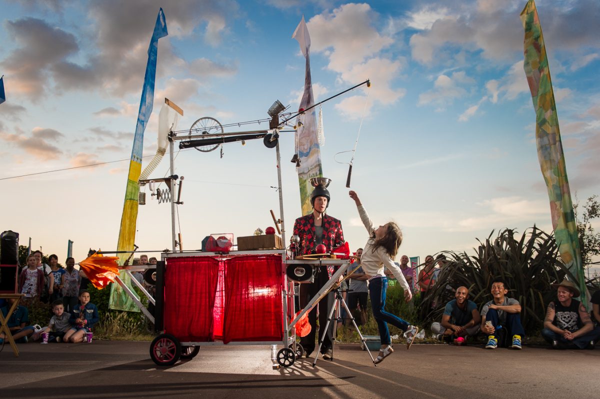 Outdoor performance featuring a performer standing inside a wheeled contraption made from metal frames, pulleys, bells and unusual mechanical objects.