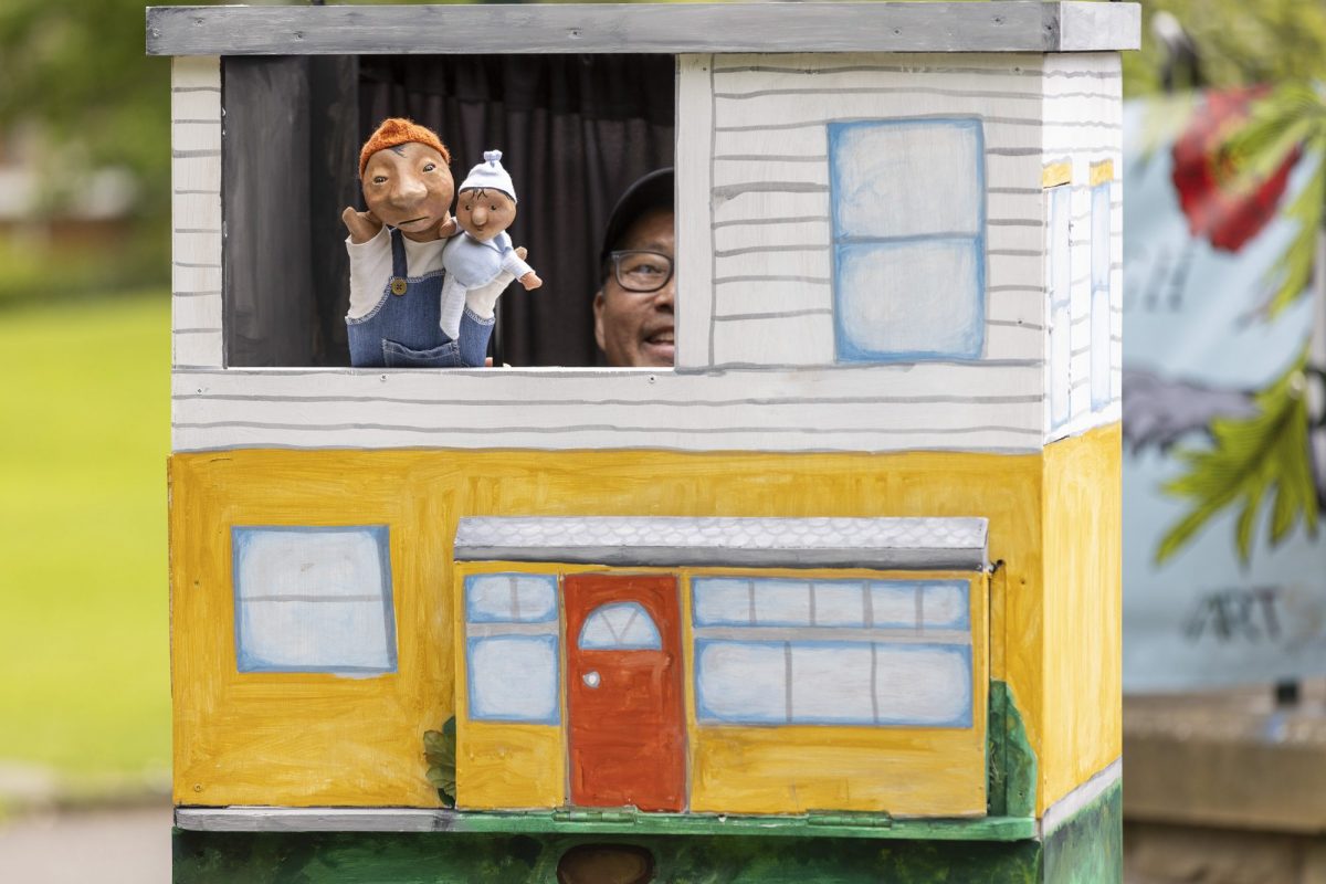 Small puppet theatre painted to look like a two-storey house with yellow walls, blue windows and a red front door. Two hand puppets, an adult and a baby, appear in an upstairs window while the puppeteer is partially visible behind them.