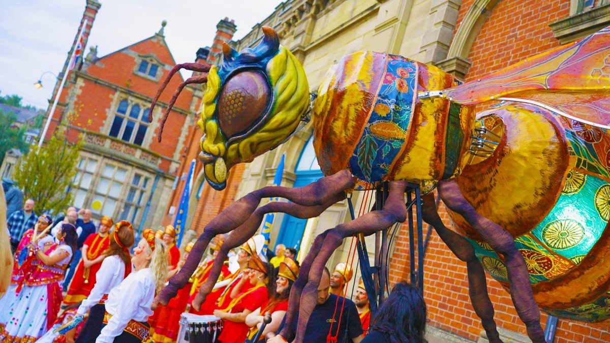 Large golden bee puppet moving through a street parade outside a historic red-brick building. The bee features colourful patterned wings and body panels, oversized eyes and long legs.