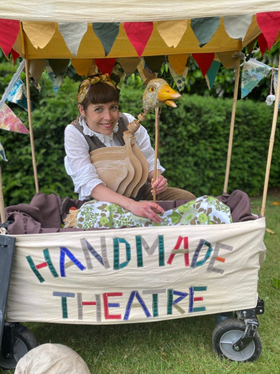 Performer seated inside a small mobile puppet theatre decorated with colourful bunting and a fabric sign reading “Handmade Theatre”. She operates a bird puppet with a long neck and beak while smiling at the audience.