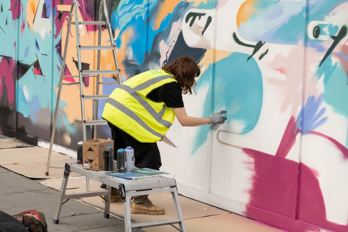 Natalie Coe, wearing a high-visibility vest, spray-paints colourful abstract forms onto a large temporary wall beside a ladder and spray cans.