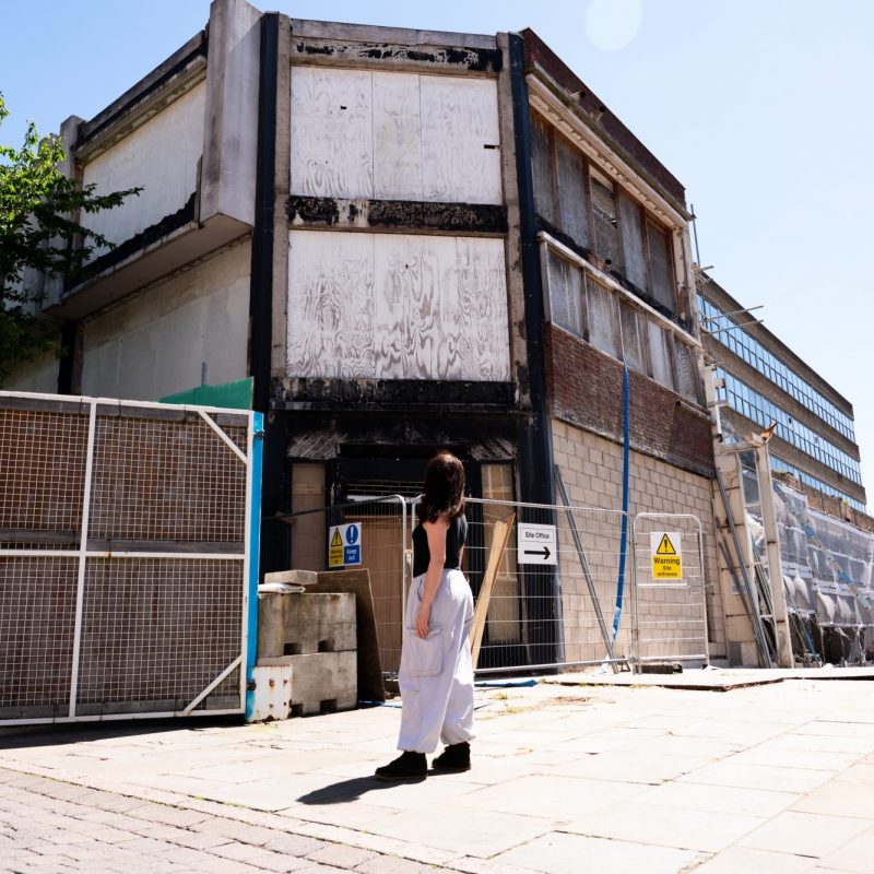 View of the boarded-up Severn’s House building in Nottingham’s Broad Marsh area, photographed before artwork begins. A person stands looking up at the building behind temporary construction fencing.
