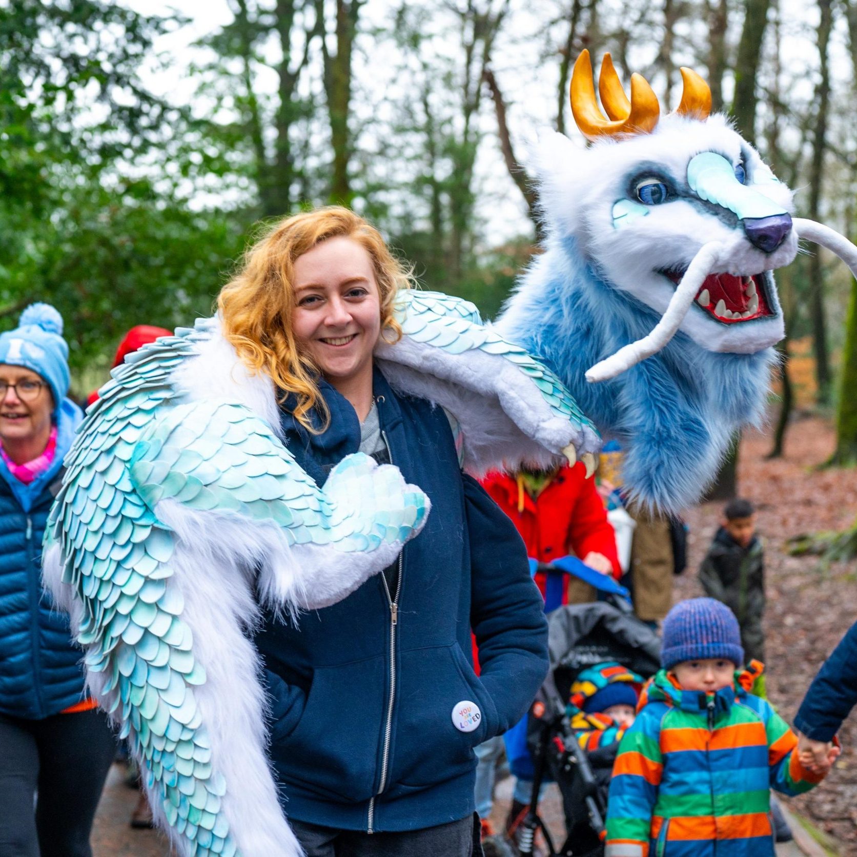 A performer carries a large blue and white dragon puppet through a woodland trail as families follow behind. The dragon has golden horns, long whiskers and feather-like scales, while children and adults in winter clothing walk alongside.