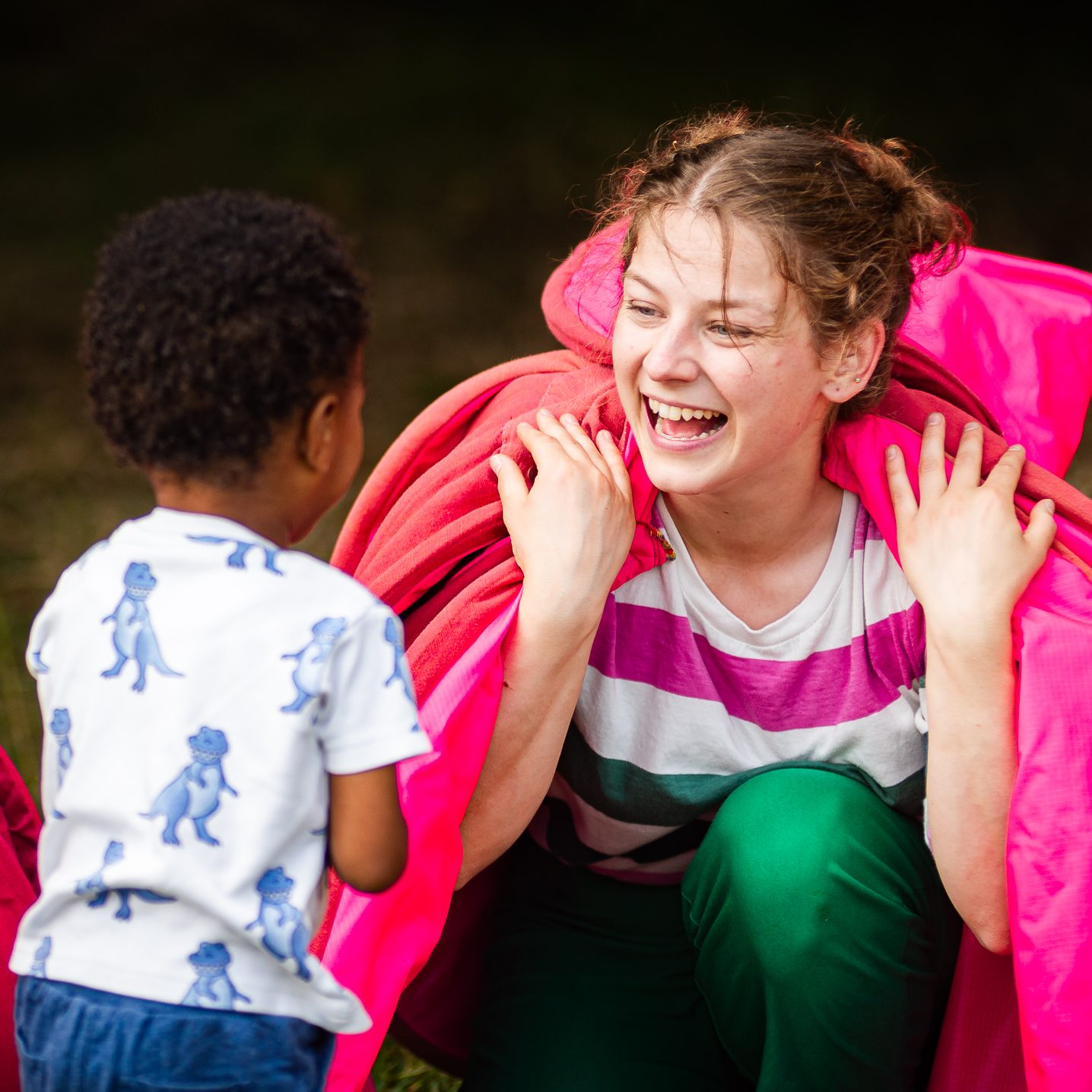 A young child smiles at a performer crouching down to their eye level during an outdoor play session.