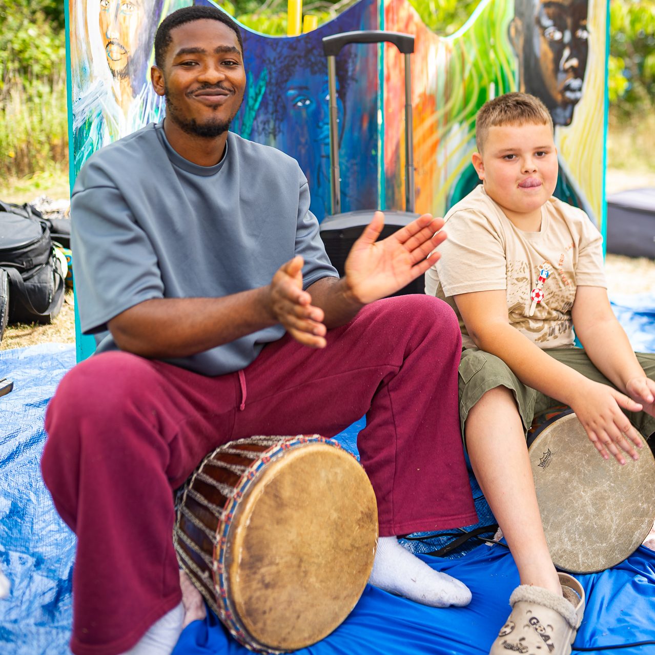 Two people sit under a pop-up gazebo playing hand drums together on a blue groundsheet.