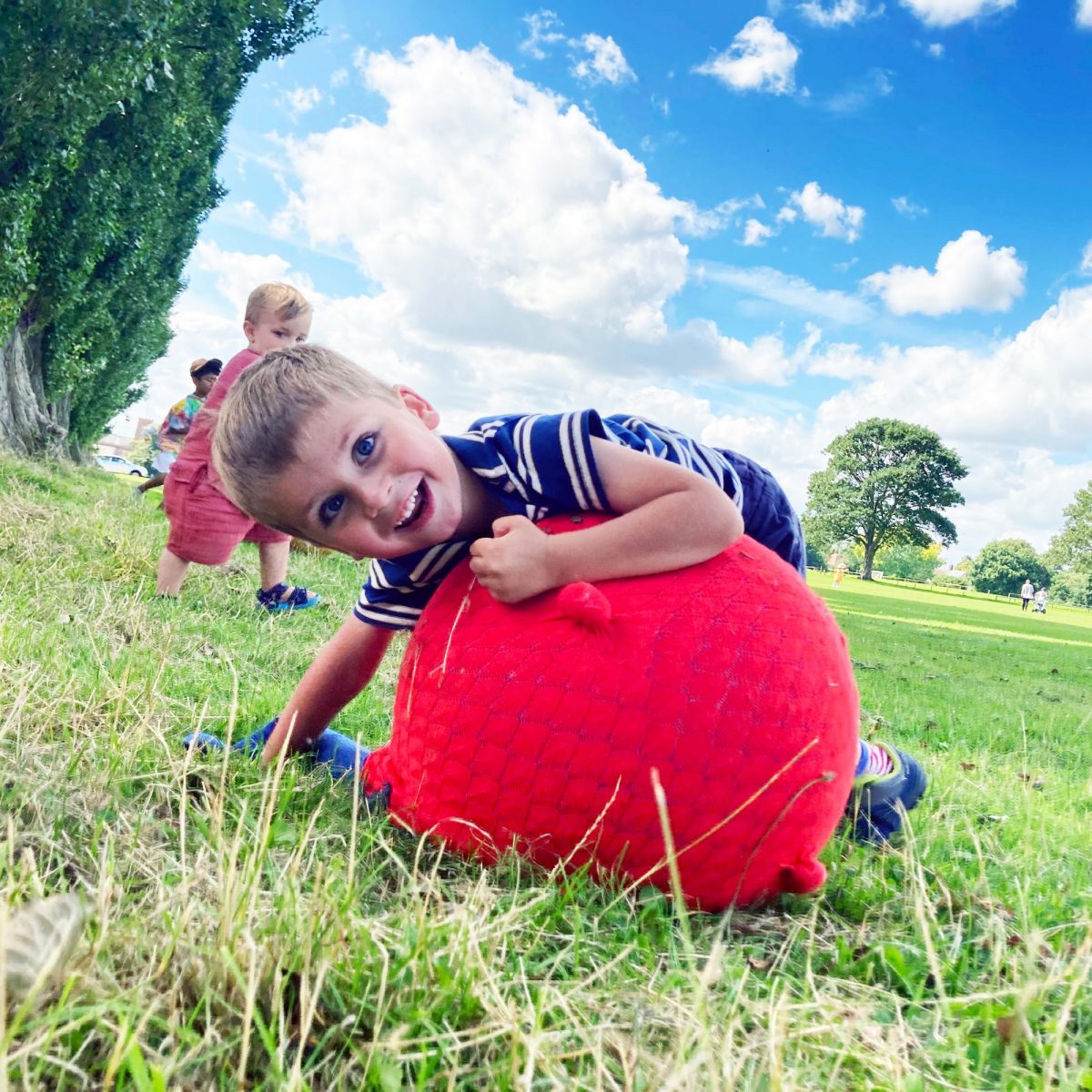 A young child smiles at the camera while hugging a large red soft play ball in a grassy park.