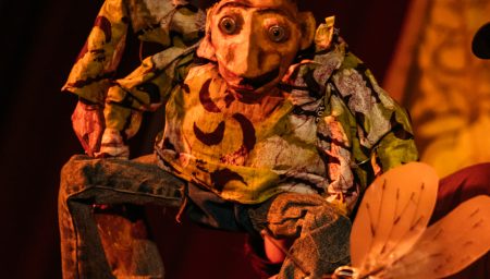 A colourful puppet of Anansi sits on stage, looking towards a large insect puppet in the foreground.