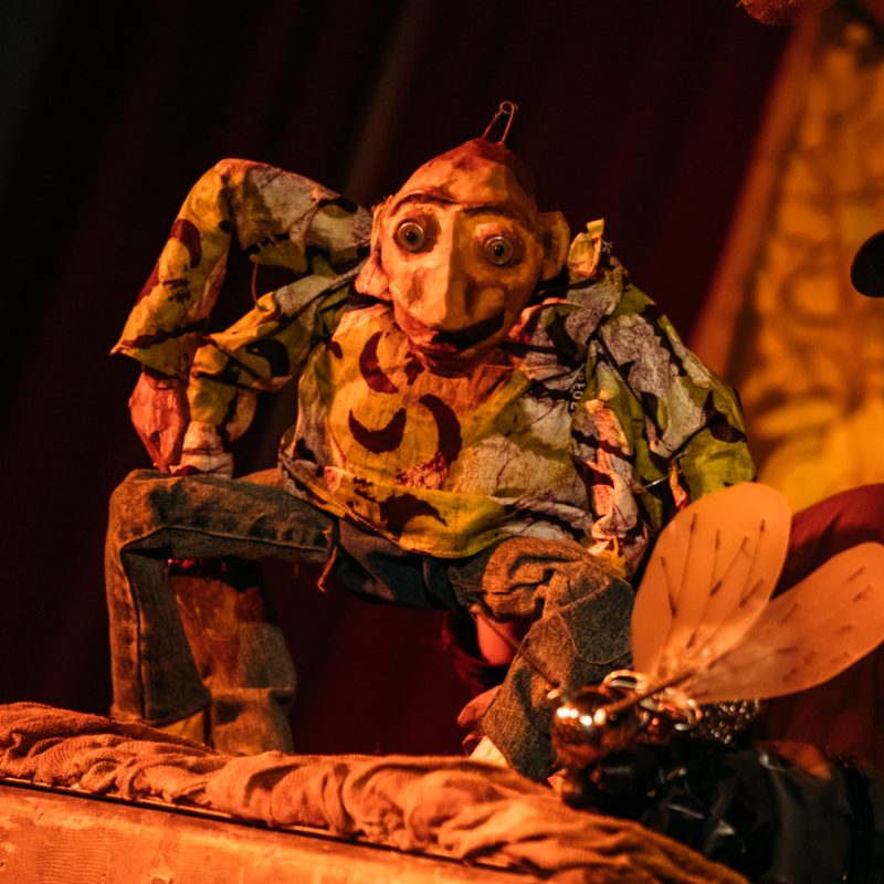 A colourful puppet of Anansi sits on stage, looking towards a large insect puppet in the foreground.