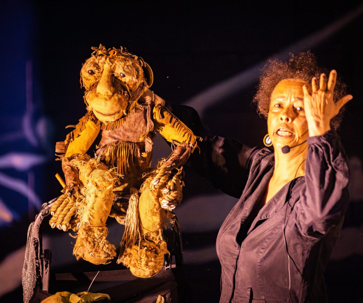 A puppeteer performs beside a large textured puppet under dramatic stage lighting.