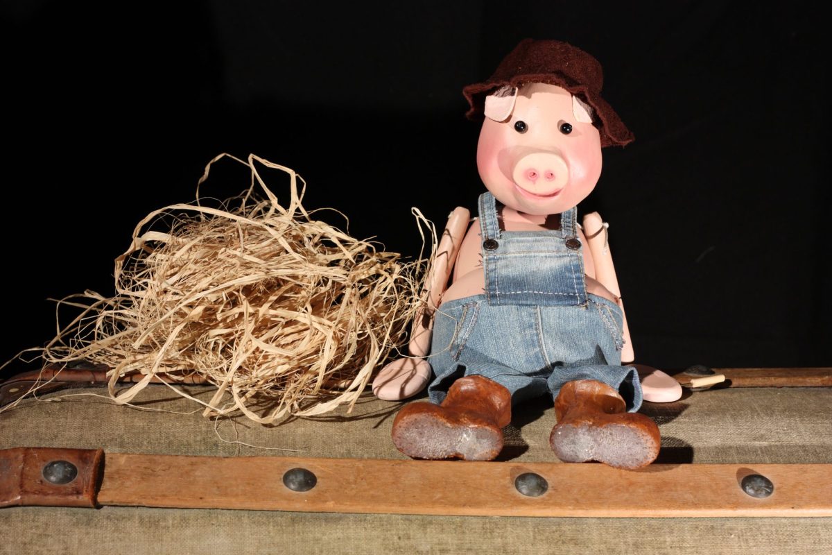 A pig puppet wearing denim dungarees, a brown hat and boots sits beside a pile of straw against a black background.